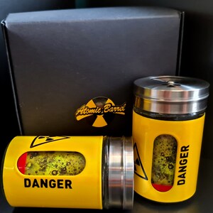 May include: Two yellow and black spice jars with silver lids. The jars have the words "DANGER" and "Atomic Barrel" printed on them.