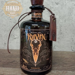 May include: A dark brown glass bottle with a black label featuring a raven skull graphic and the text "Oldschool Corps The Raven Sol". The bottle has a brown leather strap with a metal tag and a brown wooden stopper. The label also includes the text "Handcrafted Made by Lostplace Store" and "500ml".