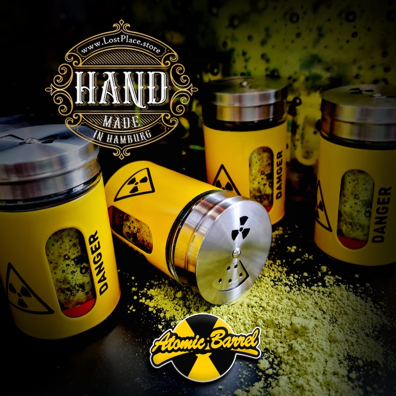 May include: Four yellow metal spice shakers with a black and yellow radioactive symbol and the word "DANGER" printed on them. The shakers are on a black surface with green powder scattered around them. The text "HAND MADE IN HAMBURG" and "www.LostPlace.store" are visible in the background. The text "Atomic Barrel" is visible in the foreground.