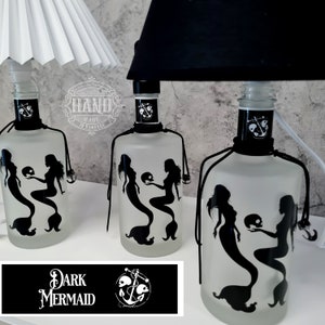 Op de afbeelding: Drie matglazen flessen met zwarte silhouetten van zeemeerminnen die schedels vasthouden. De flessen zijn versierd met zwart touw en een anker. De tekst "DARK MERMAID" staat op een zwart label met een wit anker en schedels.