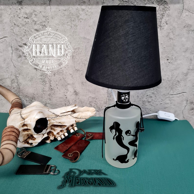 Op de afbeelding: Een zwarte lampenkap met een matglazen flesbasis met een silhouet van twee zeemeerminnen. De flesbasis heeft een zwart snoer en een witte schakelaar. De lamp staat op een turquoise oppervlak met een koeien schedel en leren sleutelhangers.