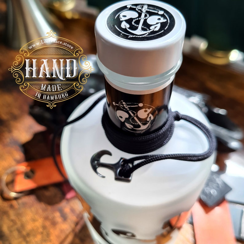 Op de afbeelding: Een witte fles met een zwart-wit schedel- en ankerontwerp. De fles is vastgebonden met een zwart koord en heeft een witte dop met een zwart schedel- en ankerontwerp. De fles is gelabeld met "HAND MADE IN HAMBURG" en "www.Lostface.store".