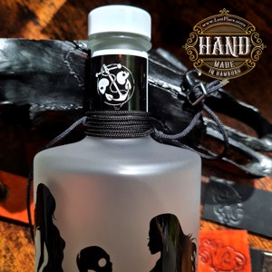 Op de afbeelding: Een frosted glazen fles met een zwart etiket met een anker en de tekst "HAND MADE IN HAMBURG". De fles is vastgebonden met zwart touw en heeft een witte dop. De fles is gedeeltelijk bedekt door een zwart lederen voorwerp.