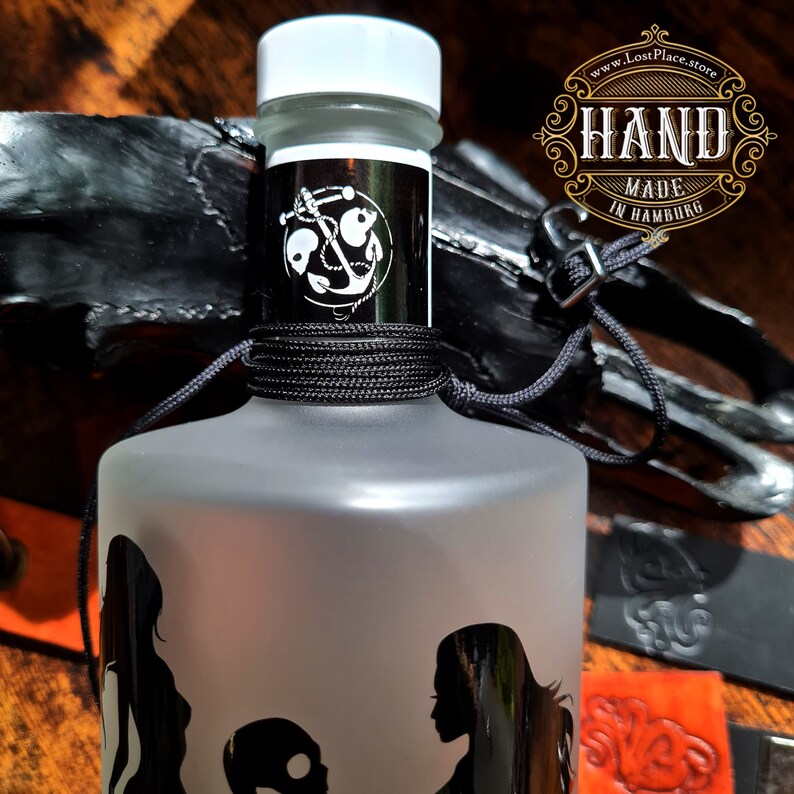 Op de afbeelding: Een frosted glazen fles met een zwart etiket met een anker en de tekst "HAND MADE IN HAMBURG". De fles is vastgebonden met zwart touw en heeft een witte dop. De fles is gedeeltelijk bedekt door een zwart lederen voorwerp.