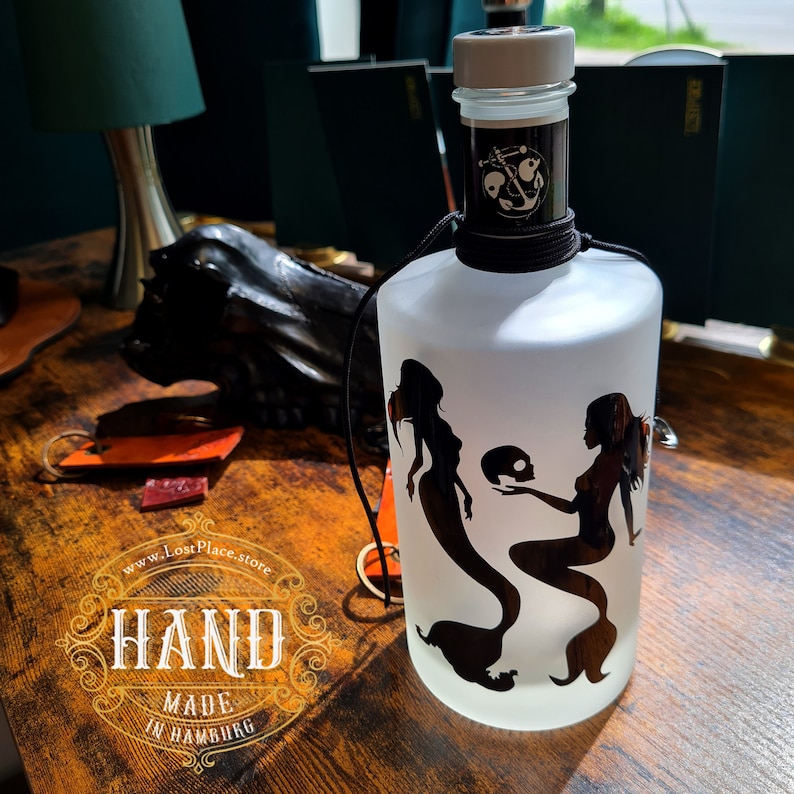 Op de afbeelding: Een frosted glazen fles met een zwart-wit silhouet van twee zeemeerminnen die een schedel vasthouden. De fles heeft een zwart koord om de nek gebonden en een zwarte dop met een ankersymbool. De fles is gelabeld met "www.LostPlace.store" en "HAND MADE IN HAMBURG".
