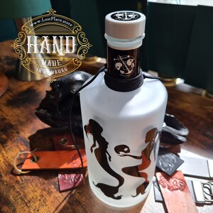 Op de afbeelding: Een gematteerde glazen fles met een zwart-wit ontwerp van een zeemeermin. De fles heeft een zwart koord om de hals gebonden en een wit deksel met een ankerontwerp. De fles is gelabeld met "Hand Made in Hamburg".