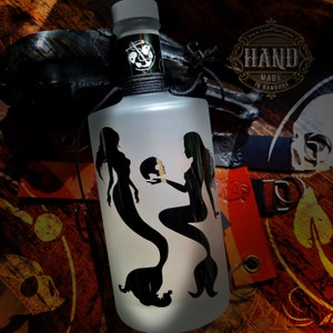 Op de afbeelding: Een frosted glazen fles met een zwarte dop en een zwart lint. De fles heeft een silhouetontwerp van twee zeemeerminnen met een schedel ertussen. De fles is gelabeld met "HAND MADE IN HAMBURG" en "www.LostPlace.store".