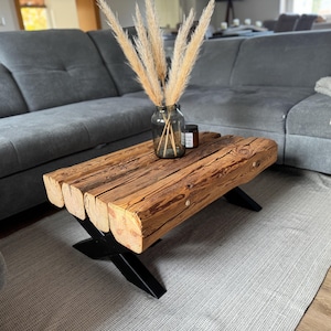 Puede incluir: Mesa de centro rústica de madera reciclada, con acabado de vetas naturales. La mesa se apoya en patas de metal negro. Un jarrón de cristal con hierba de la pampa seca y una pequeña vela encima. La mesa mide aproximadamente 122 cm de largo.