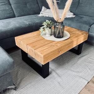 Könnte beinhalten: Rechteckiger Couchtisch aus Holz mit natürlicher Oberfläche, getragen von schwarzen Metallbeinen. Der Tisch steht auf einem grauen Teppich, im Hintergrund ein graues Sofa. Eine Vase mit dekorativen Pflanzen steht auf dem Tisch.