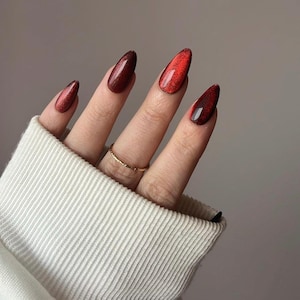 Puede incluir: Primer plano de una mano con uñas largas y ovaladas pintadas en degradado rojo y burdeos. Las uñas tienen un acabado brillante y un efecto ojo de gato. Un anillo dorado adorna el dedo anular. La mano emerge de un suéter blanco.