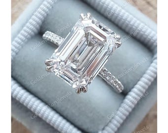 以下が含まれることがあります： 大きな透明なエメラルドカットのダイヤモンドがついたシルバーの婚約指輪。リングは、バンドに小さなダイヤモンドが敷き詰められたパヴェセッティングです。