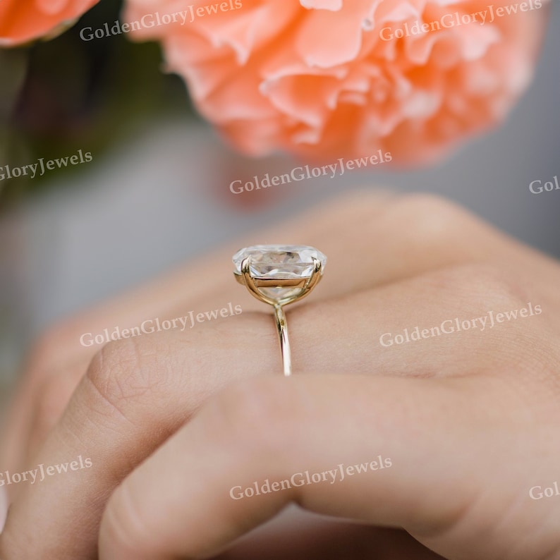 Puede incluir: Un anillo de oro con una gran piedra preciosa ovalada transparente. El anillo est&aacute; en un dedo y el fondo est&aacute; desenfocado.