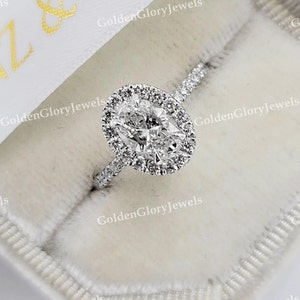Op de afbeelding: Een zilveren ring met een grote ovale diamant gezet in een halo van kleinere diamanten. De ring ligt in een witte fluwelen ringdoos.