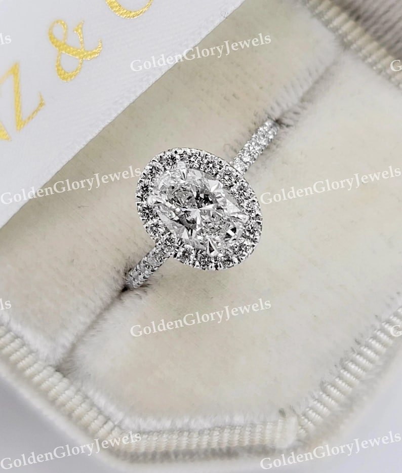 Op de afbeelding: Een zilveren ring met een grote ovale diamant gezet in een halo van kleinere diamanten. De ring ligt in een witte fluwelen ringdoos.
