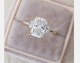 May include: A gold engagement ring with a large, square-shaped diamond center stone and two smaller, pear-shaped diamonds on either side. The ring is in a velvet ring box.