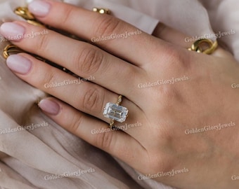 Puede incluir: Un anillo de oro con una gran piedra preciosa transparente de talla esmeralda. El anillo está en un dedo.