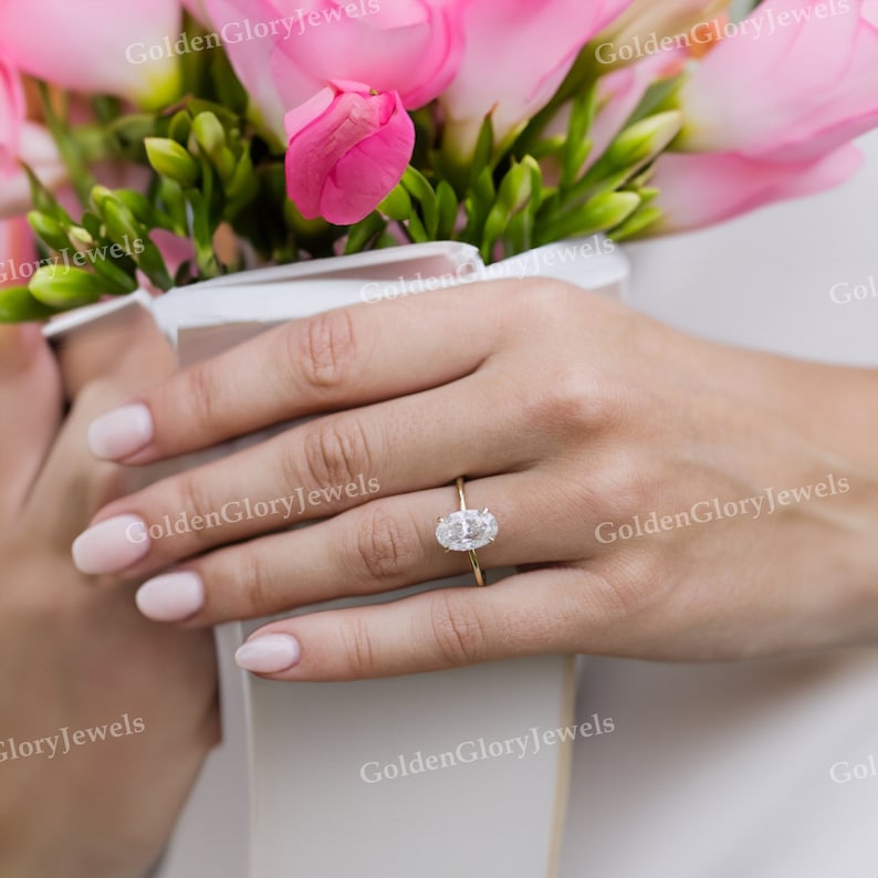 Puede incluir: Primer plano de una mano con un anillo de oro con un gran diamante de forma ovalada. El anillo est&aacute; engastado en una banda sencilla.