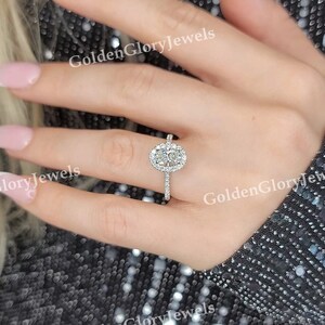 Op de afbeelding: Een close-up van een hand met een zilveren ring met een ovale diamant omringd door kleinere diamanten. De ring is gezet op een dunne band.