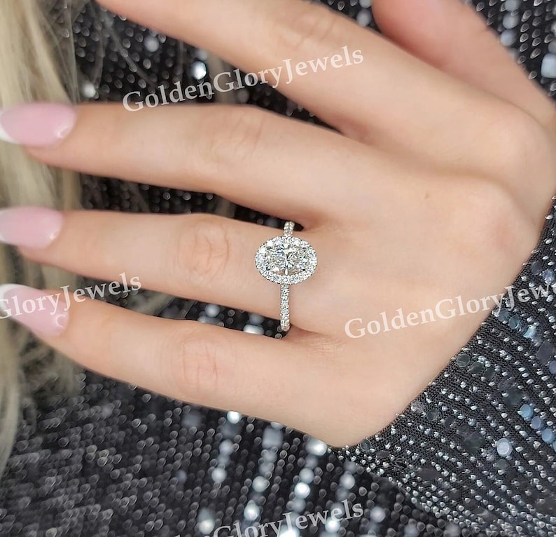Op de afbeelding: Een close-up van een hand met een zilveren ring met een ovale diamant omringd door kleinere diamanten. De ring is gezet op een dunne band.