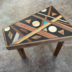 May include: A triangular wooden table with a geometric design featuring various wood tones, black, red, and blue accents. The table has three legs and is made of solid wood.