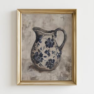 May include: A framed painting of a ceramic pitcher with a handle. The pitcher is decorated with blue floral patterns on a cream background. The frame is gold-colored, and the background is a neutral gray.