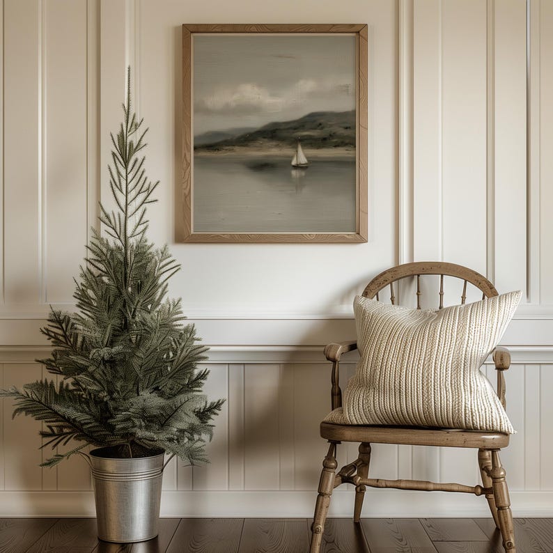 May include: A decorative interior scene featuring a small artificial evergreen tree in a silver bucket, a framed landscape painting of a sailboat on water, and a wooden chair with a cream-colored pillow. The wall is white with vertical paneling.