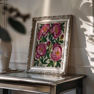 May include: A framed floral painting featuring pink peonies with yellow centers and green leaves. The artwork is set in a silver ornate frame, resting on a dark-colored table. The painting is illuminated by natural light.