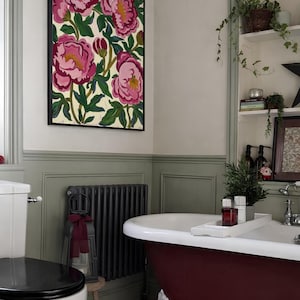 May include: A bathroom scene featuring a framed floral artwork with pink peonies. A burgundy clawfoot bathtub is in the foreground, complemented by a black radiator and a white toilet. The walls are painted in neutral tones.