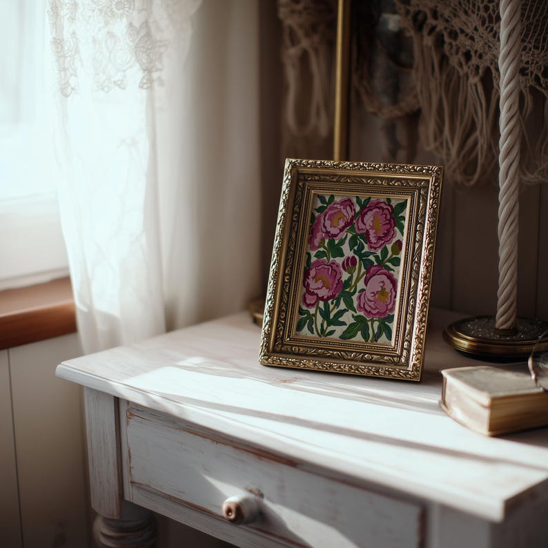 May include: A gold framed picture with a floral design featuring pink flowers and green leaves. The picture is displayed on a white painted wooden table. A small book and lamp are also on the table, near a window with a white curtain.