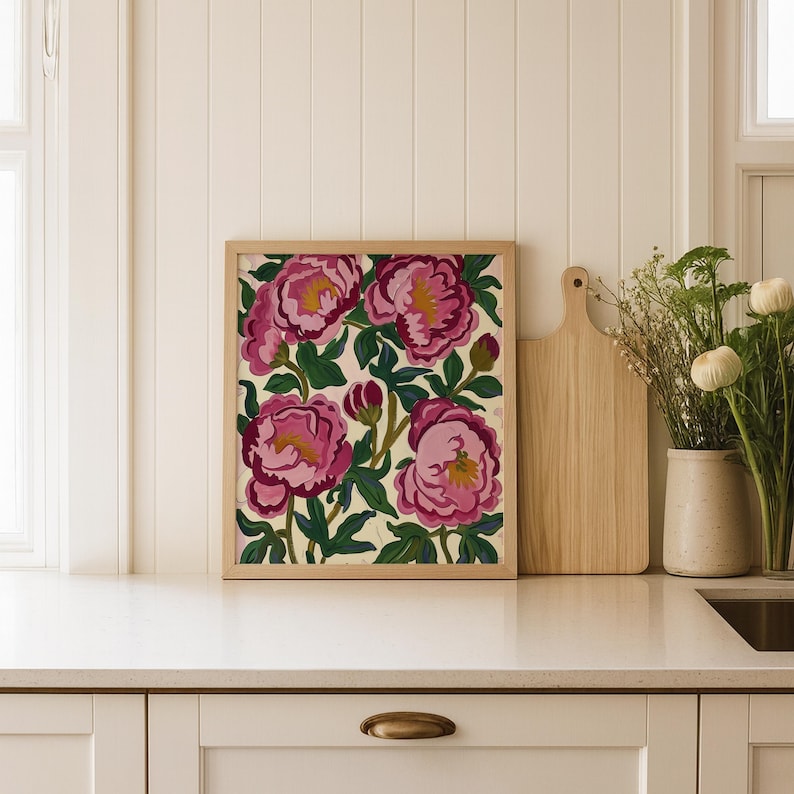 May include: A framed floral print featuring large pink peonies with yellow centers and green leaves. The artwork is in a light wood frame and rests on a white countertop. The overall theme is botanical art.