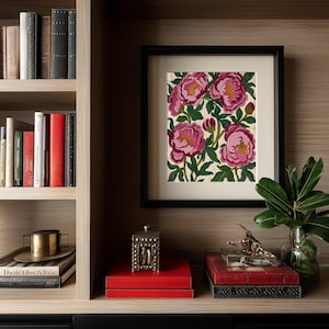 May include: A framed floral print featuring pink peonies with yellow centers and green leaves. The artwork is displayed on a shelf with books, decorative boxes, and a small gold sculpture. The frame is black.