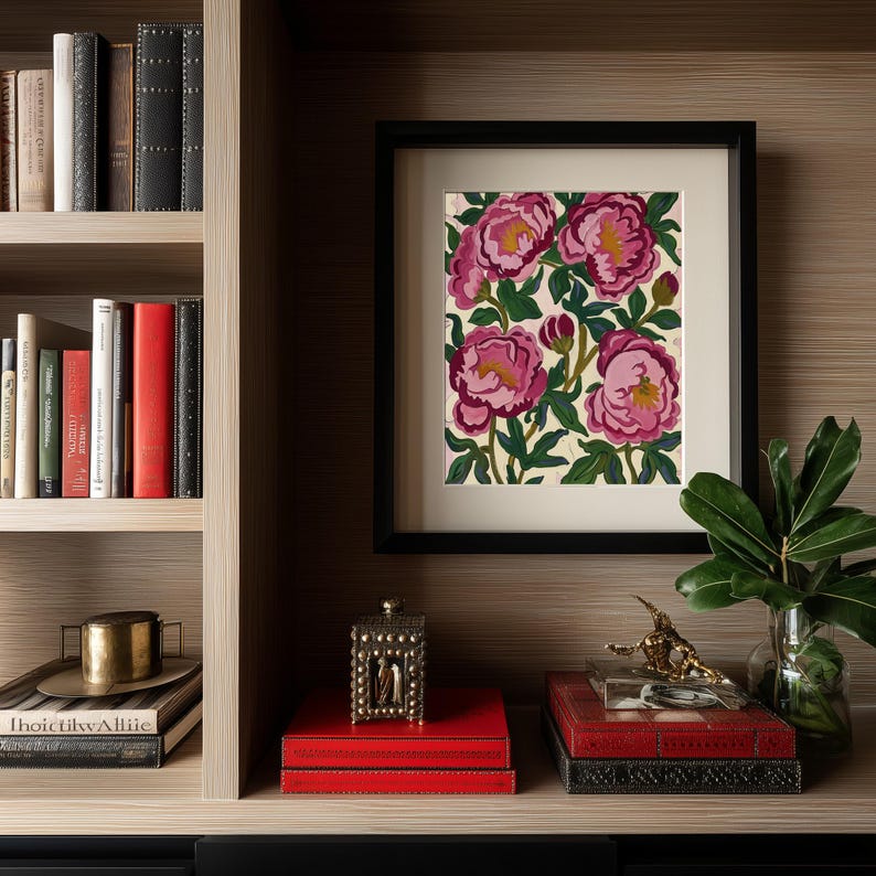 May include: A framed floral print featuring pink peonies with yellow centers and green leaves. The artwork is displayed on a shelf with books, decorative boxes, and a small gold sculpture. The frame is black.