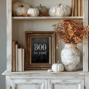 May include: A rectangular picture frame with a dark, distressed finish containing a black chalkboard insert with the white text "BOO to you!". The frame is displayed on a white shelf alongside a white ceramic vase filled with dried foliage.