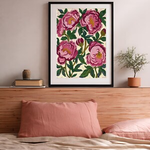 May include: Framed floral art print featuring large pink peonies with yellow centers and green leaves against a cream background. The artwork is in a black frame and is displayed above a bed with a pink pillow and a book.
