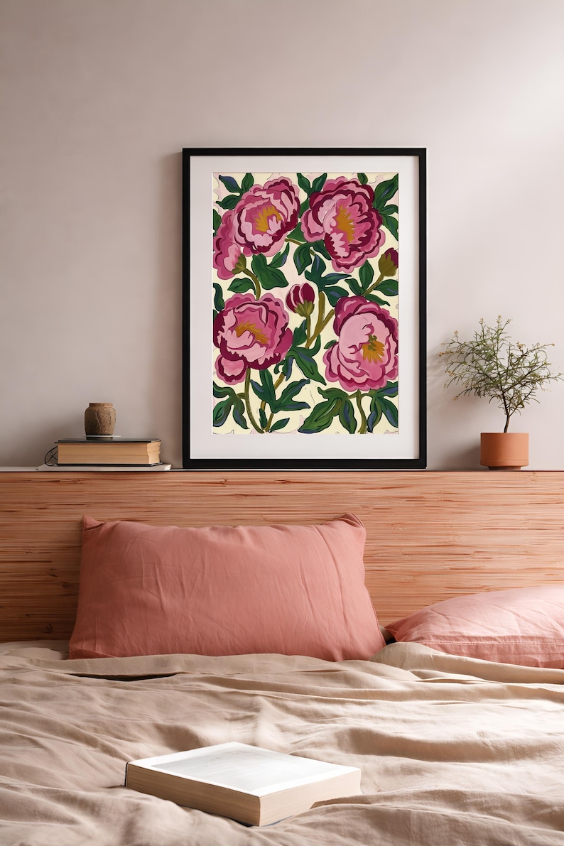 May include: Framed floral art print featuring large pink peonies with yellow centers and green leaves against a cream background. The artwork is in a black frame and is displayed above a bed with a pink pillow and a book.