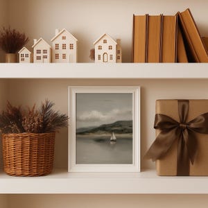 May include: A decorative shelf arrangement featuring a framed landscape painting of a sailboat on water, miniature white houses, a woven basket with dried plants, a wrapped gift with a brown ribbon, and stacked books.