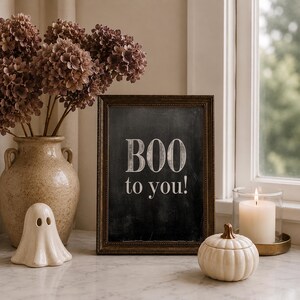 May include: A rectangular dark brown frame containing a black chalkboard sign with the white text BOO to you!. The display includes a beige ceramic vase with dried brown hydrangea flowers, a white ghost figurine, and a small white pumpkin.