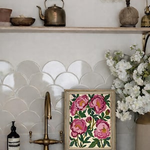 May include: A framed floral painting featuring pink peonies with yellow centers and green leaves. The artwork is displayed on a countertop with a brass faucet and a white sink. A shelf above holds a teapot and decorative items.