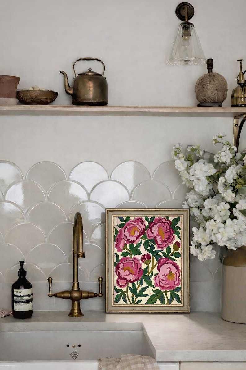 May include: A framed floral painting featuring pink peonies with yellow centers and green leaves. The artwork is displayed on a countertop with a brass faucet and a white sink. A shelf above holds a teapot and decorative items.