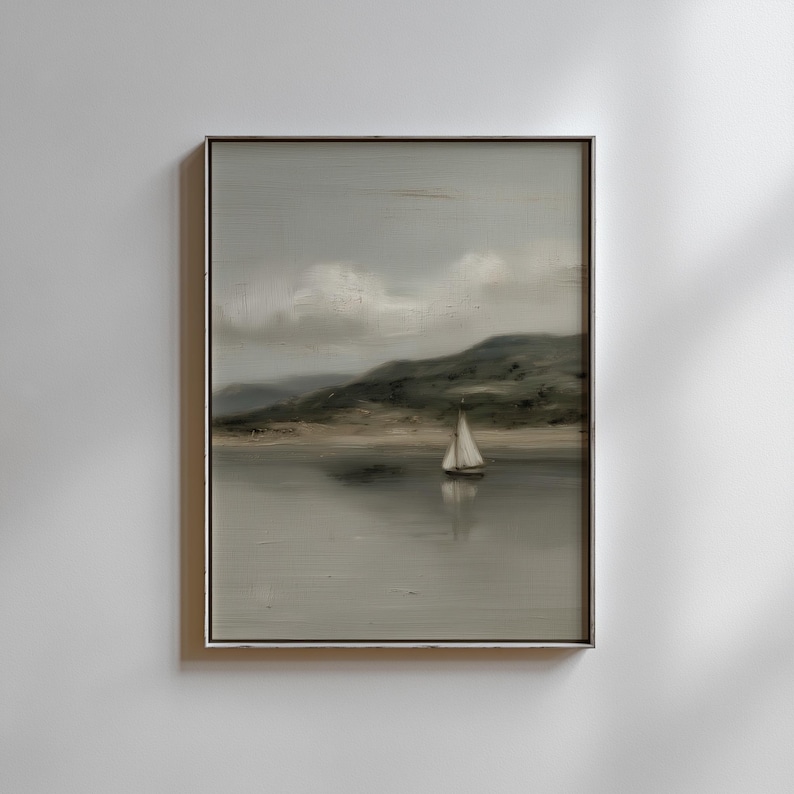 May include: A framed landscape painting depicts a sailboat on a calm lake. The artwork features a muted color palette of gray, green, and beige, with a cloudy sky and rolling hills in the background. The painting is in a silver frame.