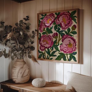 May include: A framed floral artwork featuring large pink peonies with yellow centers and green leaves. The artwork is in a light wood frame and hangs on a white paneled wall. A woven vase with greenery and a small round pouf are on a wooden surface.