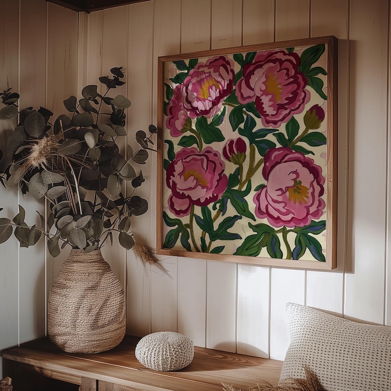 May include: A framed floral artwork featuring large pink peonies with yellow centers and green leaves. The artwork is in a light wood frame and hangs on a white paneled wall. A woven vase with greenery and a small round pouf are on a wooden surface.