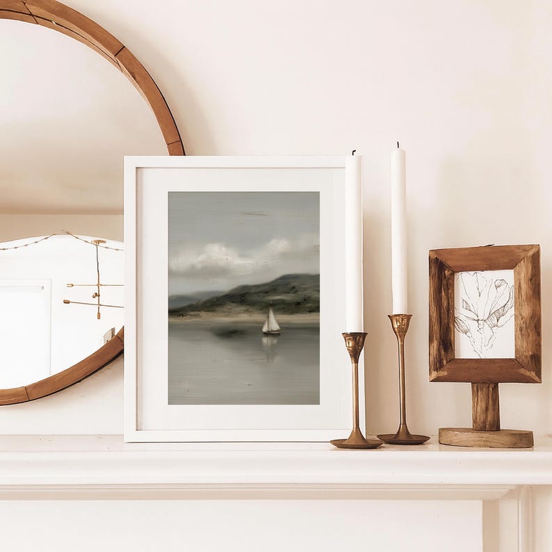 May include: A framed landscape print of a sailboat on the water, with a white frame. Two brass candle holders with white candles and a small wooden framed drawing are on a white surface.