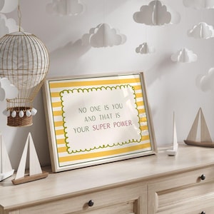 May include: A rectangular framed print featuring a white background with horizontal yellow stripes. The center displays a scalloped-edge box containing the text: NO ONE IS YOU AND THAT IS YOUR SUPER POWER, with the word SUPER in red lettering.
