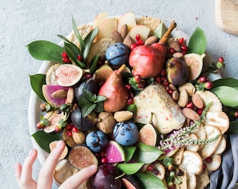 May include: A white platter filled with a variety of fruits, nuts, and cheese. The platter includes figs, pears, apples, plums, almonds, and a round wheel of cheese. The platter is surrounded by green leaves and sprigs of flowers.