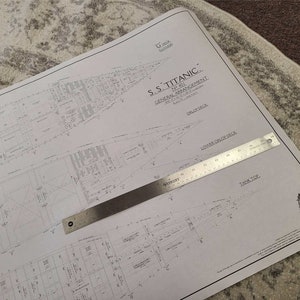 以下が含まれることがあります： S.S.タイタニック号の詳細な設計図で、建築図とテキストが記載されています。金属製の定規が紙の上に置かれ、縮尺を示しています。この文書は明るい色の表面にあり、船の一般的な配置を示しています。