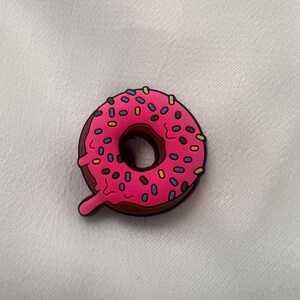 May include: A pink doughnut with brown icing and colourful sprinkles. The doughnut has a hole in the centre and a drip of icing on the side.