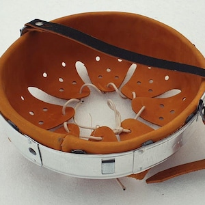 Puede incluir: Un casco antiguo con una banda de metal plateado y una correa de cuero marrón. El interior está forrado con cuero naranja y presenta fijaciones de cuerda blanca. El casco tiene varios pequeños orificios de ventilación.