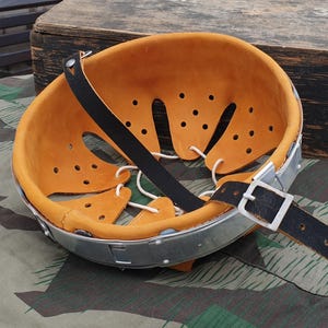 May include: A helmet liner featuring a tan leather interior with multiple ventilation holes and white cord lacing. The liner is attached to a silver-coloured metal band and includes a black leather chin strap with a metal buckle for adjustment.