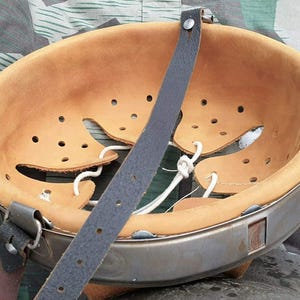 Puede incluir: Un casco militar vintage, visto desde el interior. El interior es de cuero marrón con numerosos orificios de ventilación y cordones blancos. Una correa de cuero gris oscuro está unida al borde metálico, de color plateado mate.